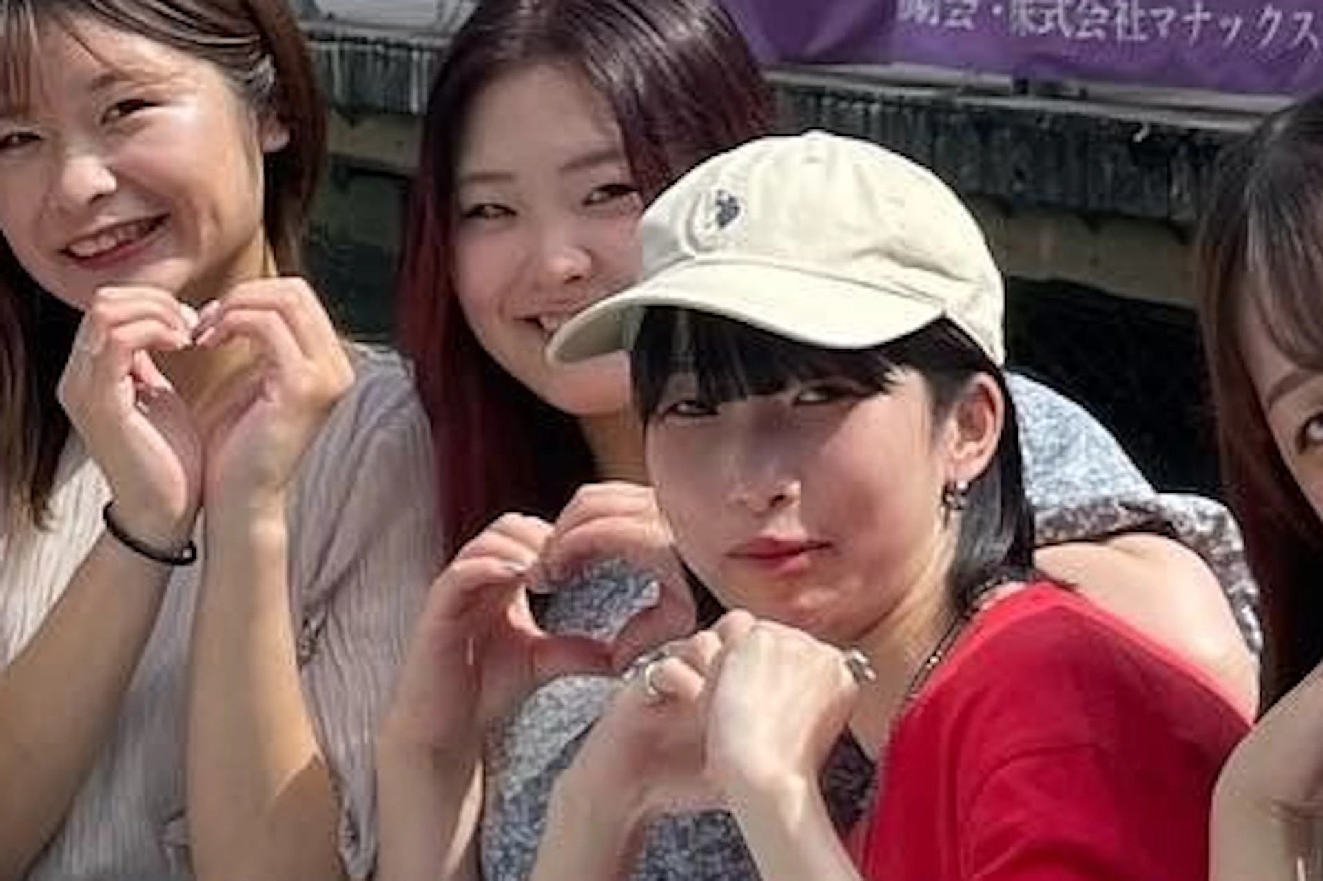 A group of seven women posing outdoors near a canal, making hand heart gestures, with colorful urban buildings in the background.