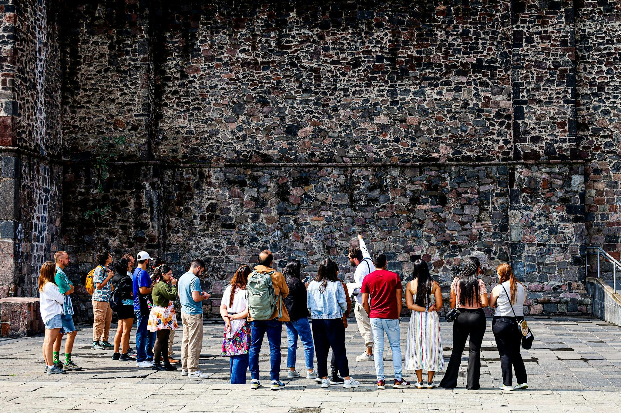 A group of people standing near an old stone wall, some facing forward and others listening to a person speaking.
