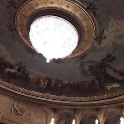 En stor ljuskrona hänger från ett utsmyckat, väggmålat tak i en överdådig teater eller konsertsal.