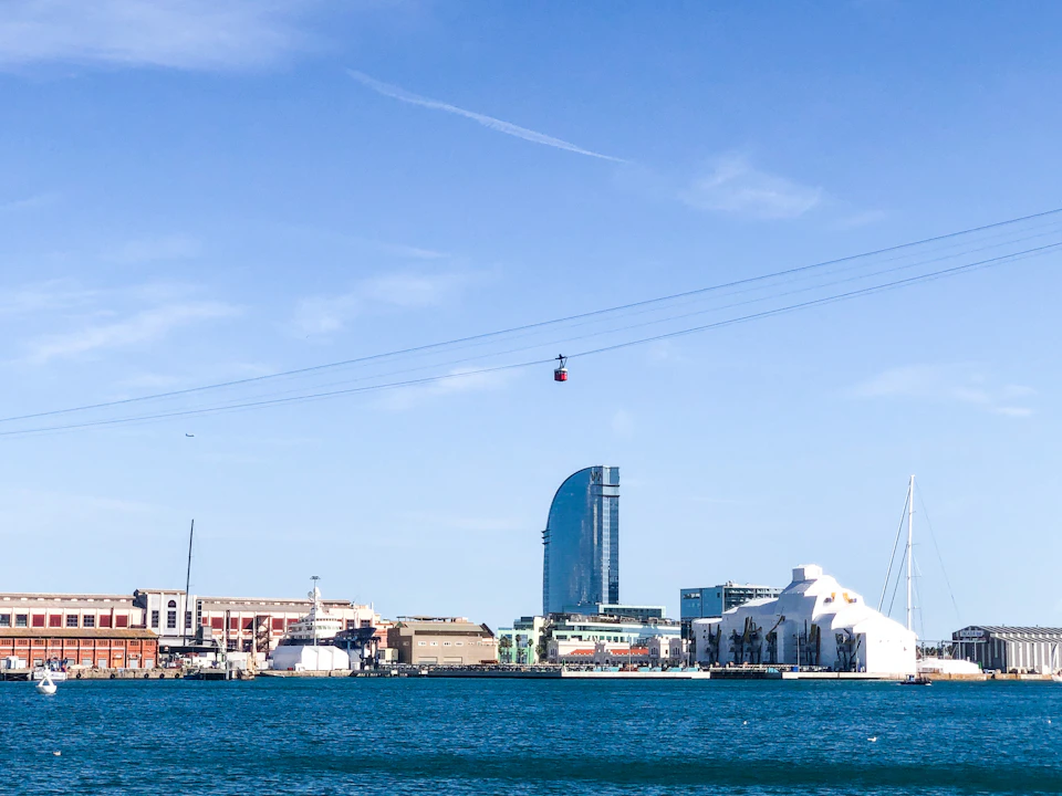 A coastal cityscape features a tall, curved skyscraper, several low-rise buildings, and a single cable car crossing a blue sky.