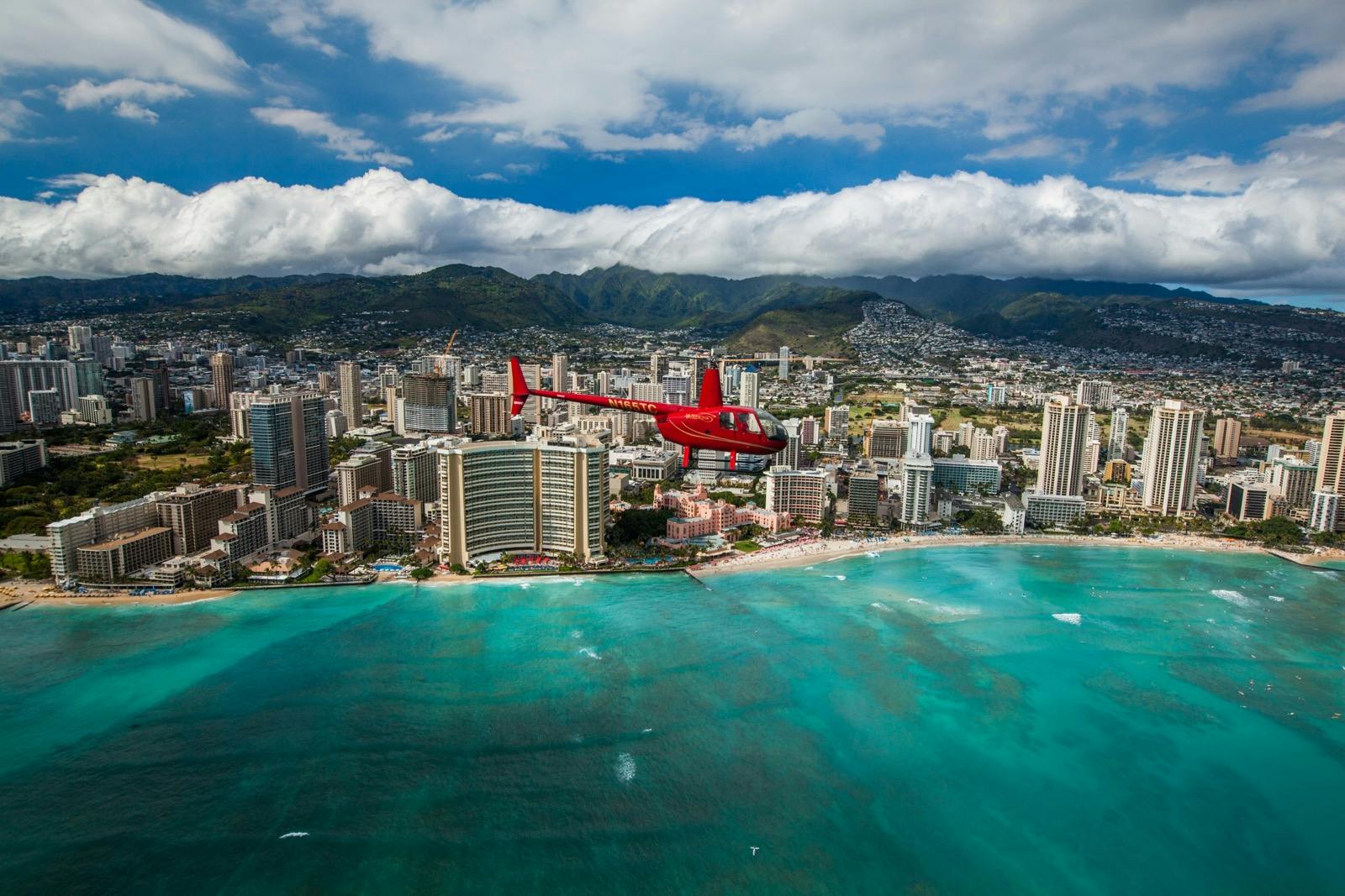 A red helicopter flying over a coastal city with tall buildings, green mountains in the background, and turquoise ocean waters below.
