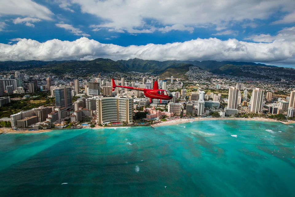A red helicopter flying over a coastal city with tall buildings, green mountains in the background, and turquoise ocean waters below.