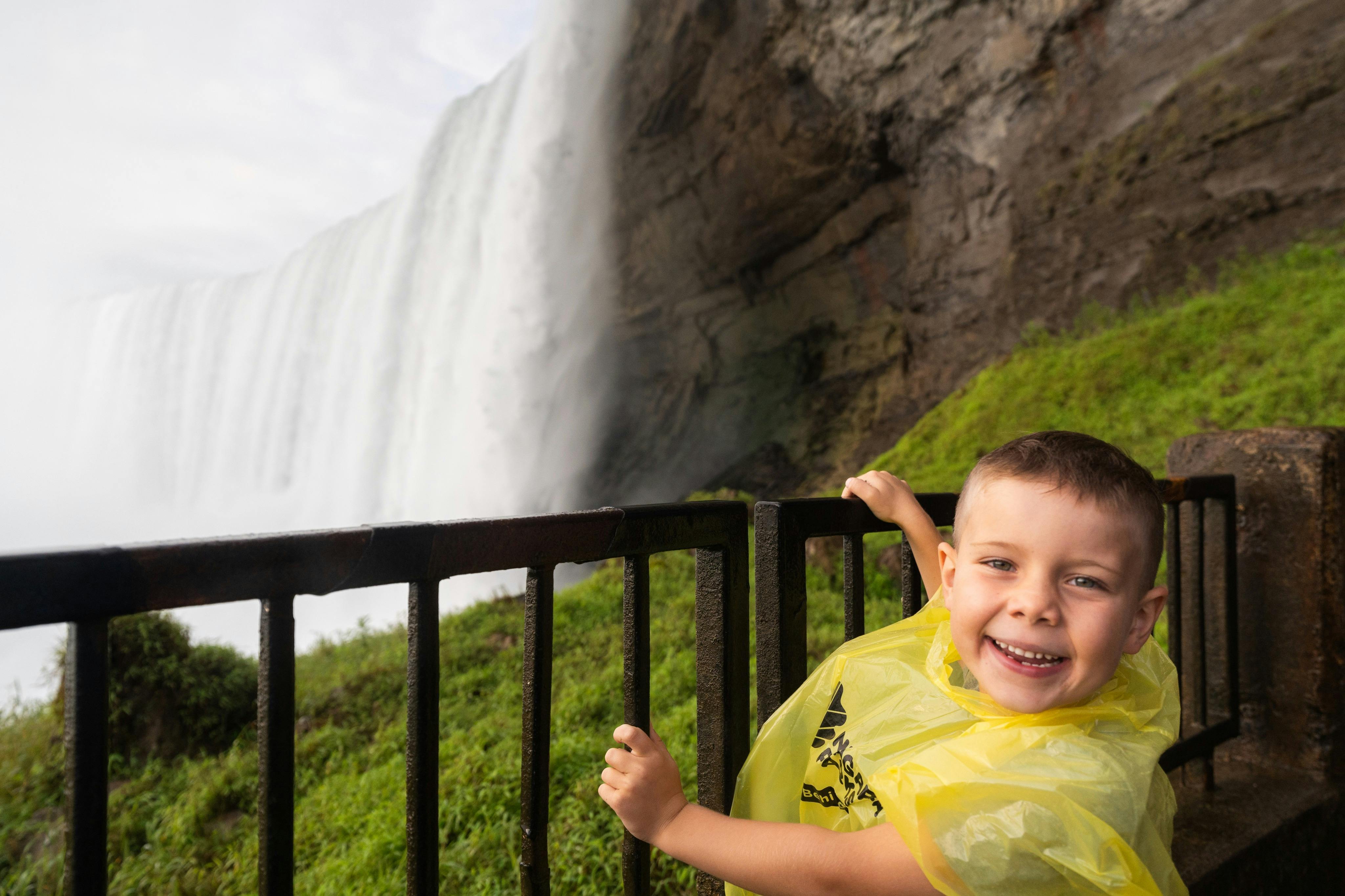 U zábradlí s vodopádem a skalnatým útesem v pozadí stojí usměvavé dítě ve žlutém dešťovém pončo.