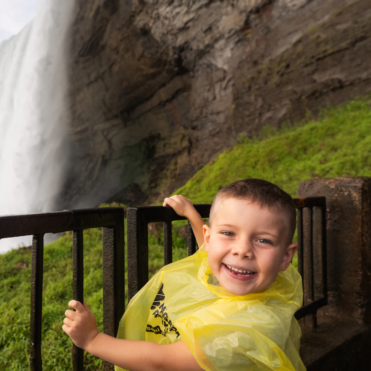 A smiling child in a yellow rain poncho stands near a railing with a waterfall and rocky cliff in the background.