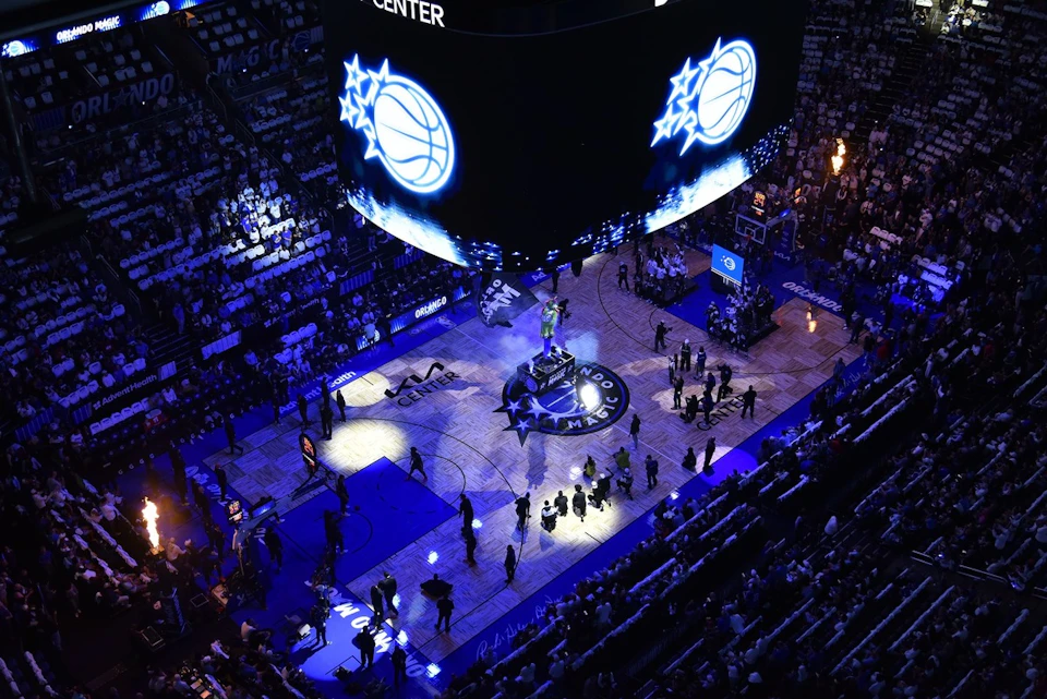 A basketball arena with a lit-up court featuring the Orlando Magic logo, surrounded by a crowd and large screens overhead.