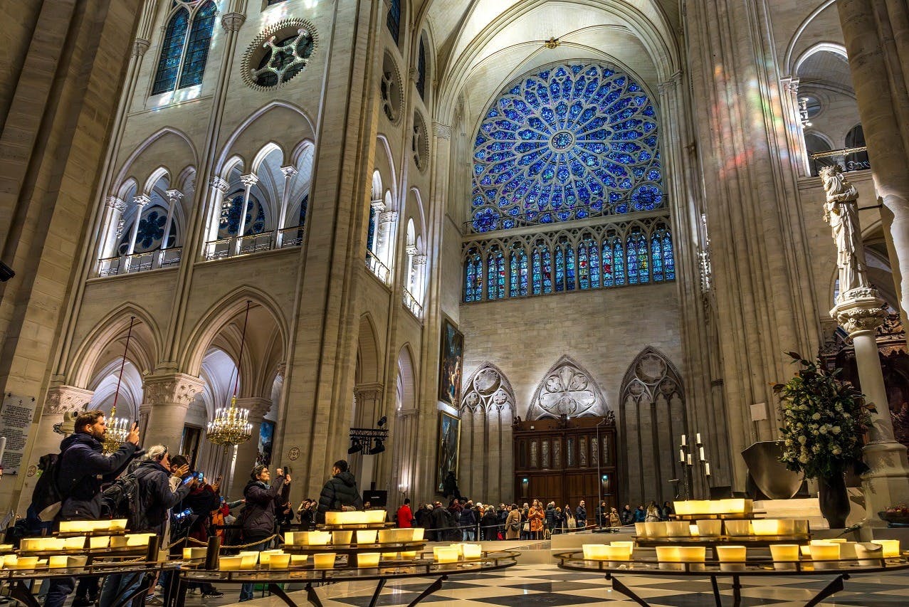 Interior of a cathedral with a large blue stained glass rose window, arched columns, and visitors taking photos near lit candles.