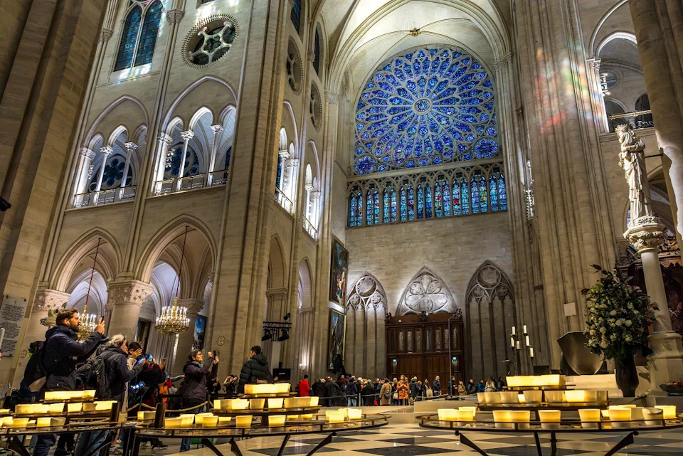 Interior of a cathedral with a large blue stained glass rose window, arched columns, and visitors taking photos near lit candles.