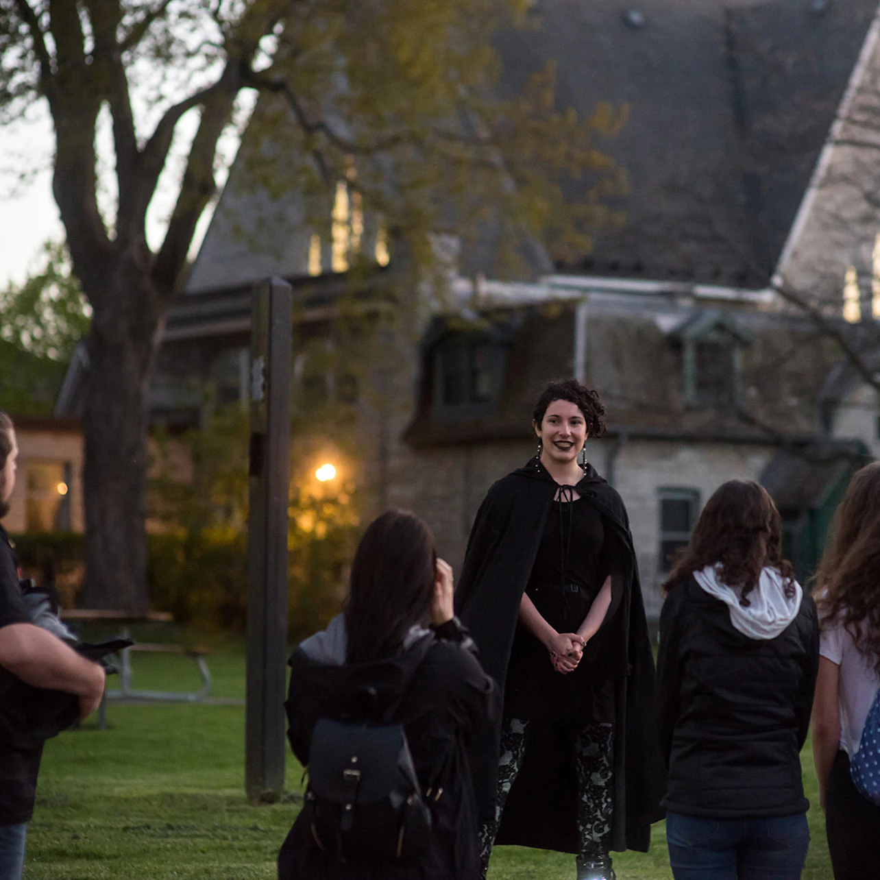 A person in a black cape speaks to a group of people on a grassy area in front of a large building at dusk.