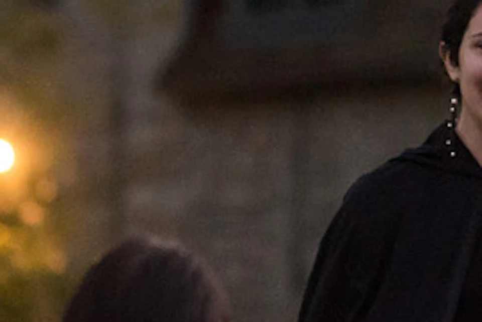 A person in a black cape speaks to a group of people on a grassy area in front of a large building at dusk.