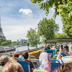 Turister på en öppen buss som tar bilder av Eiffeltornet vid floden Seine under en klarblå himmel.