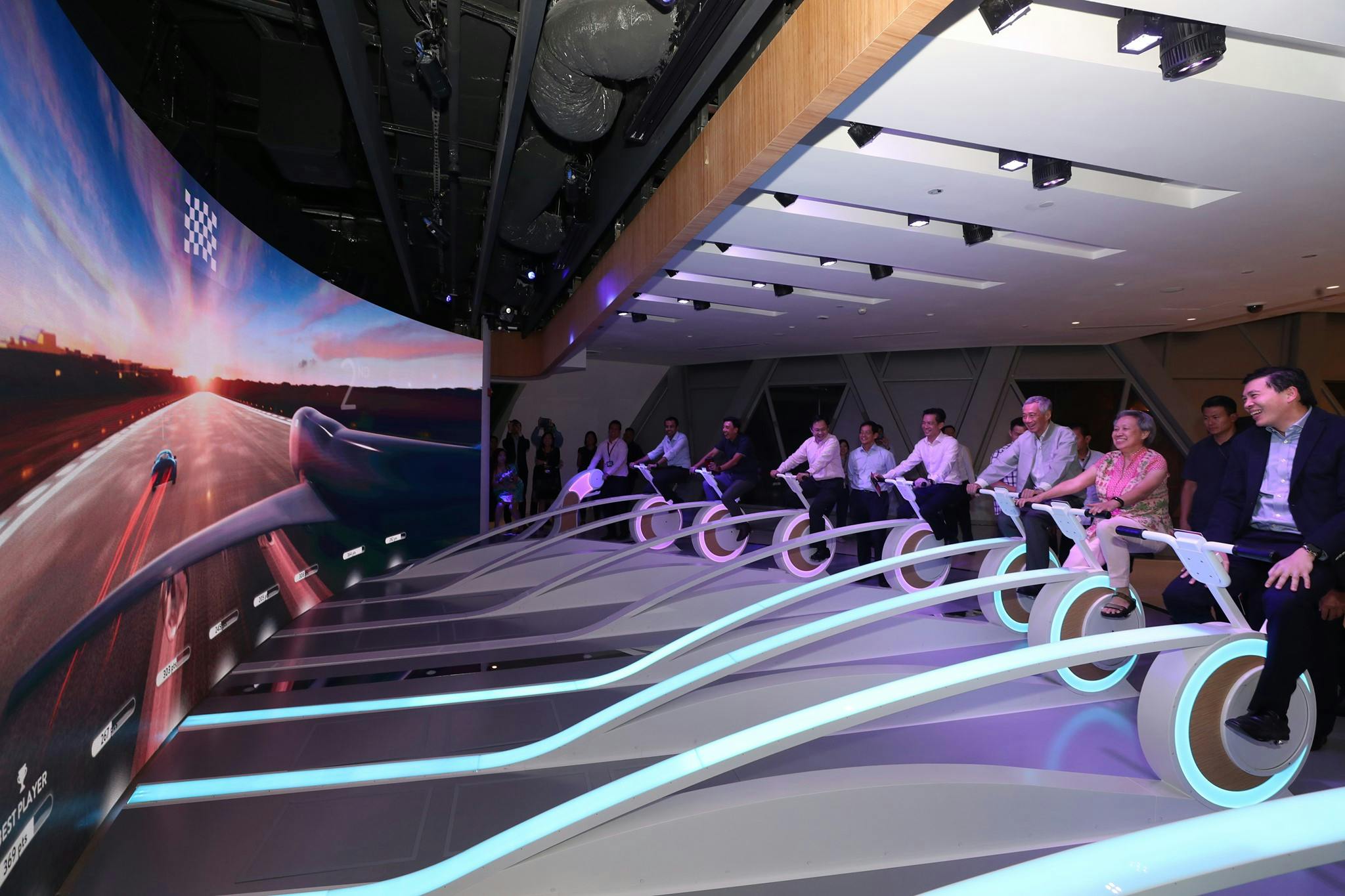 Lidé v obchodních oděvech jezdí na futuristických stacionárních kolech s osvětlenými koly uvnitř, které sleduje malé publikum.