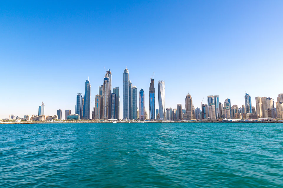 Skyline of modern skyscrapers seen across calm blue water under a clear blue sky.
