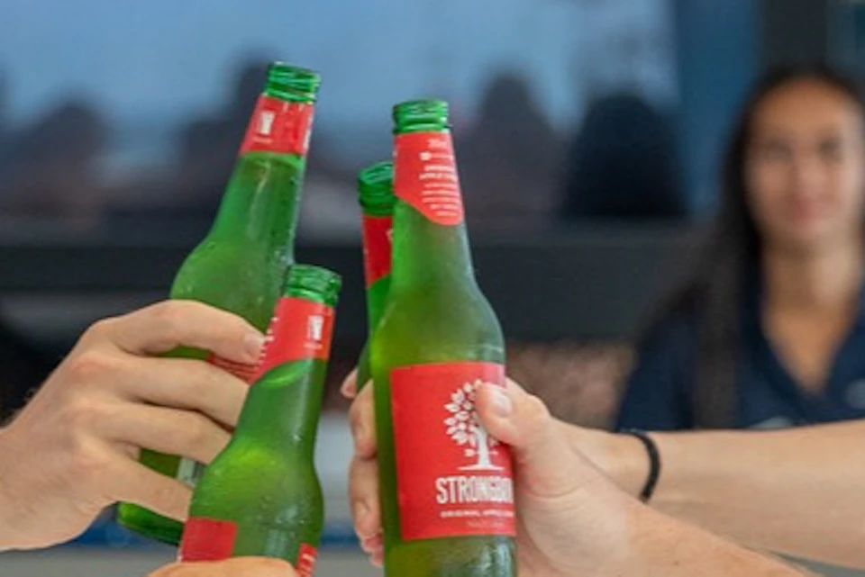 Three people clink green beer bottles while smiling at a table, with a person in the background.
