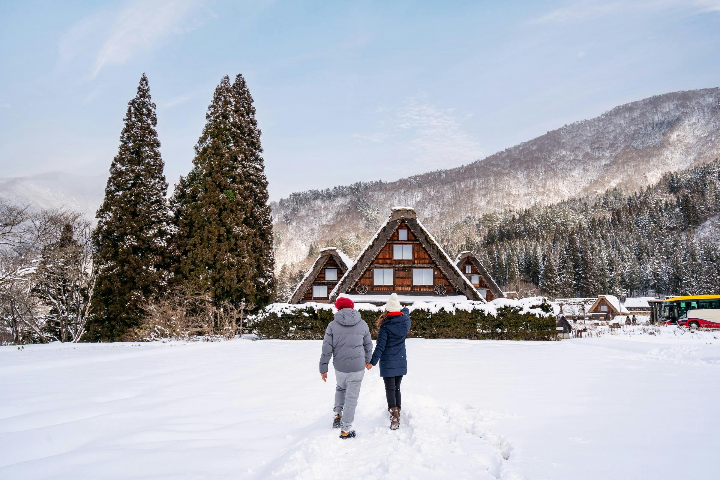Two people walking in snow towards traditional wooden houses surrounded by trees and mountains on a clear day.