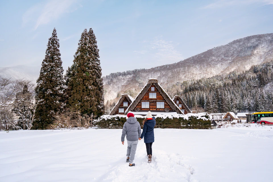 Two people walking in snow towards traditional wooden houses surrounded by trees and mountains on a clear day.