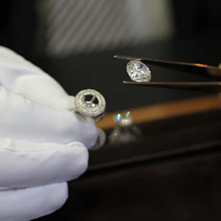 En hand i en vit handske håller en ring, medan pincett håller en lös diamant ovanför en svart yta.