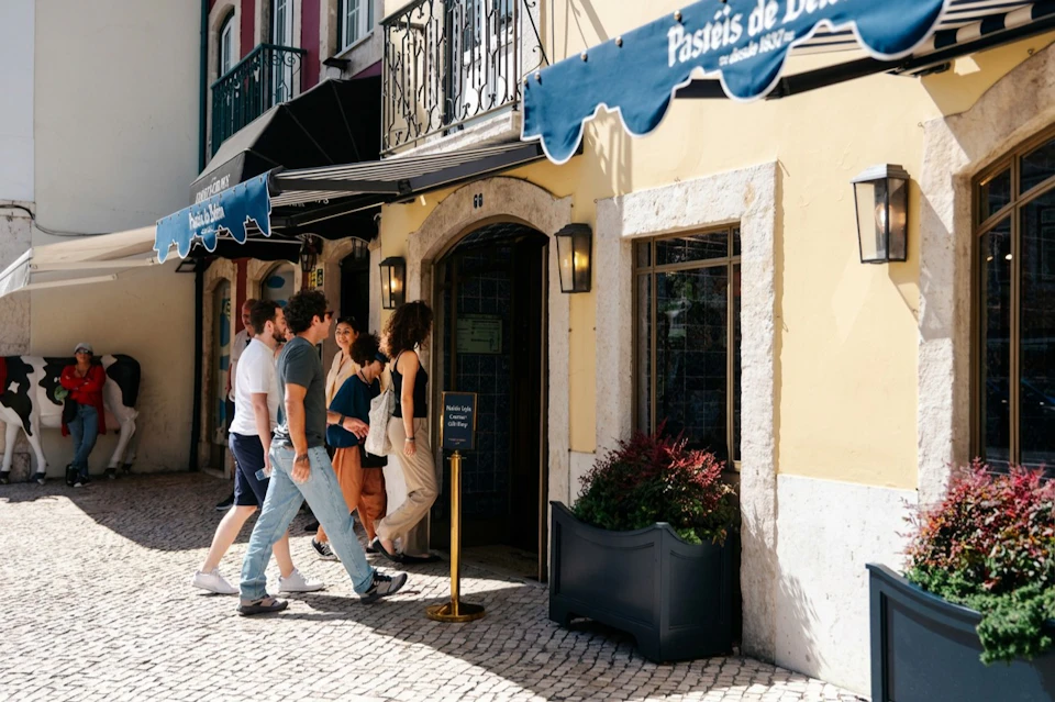 People walking into a pastry shop with a blue awning on a sunny street with cobblestone pavement.