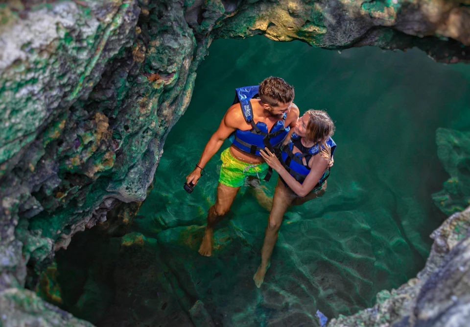Two people in blue life vests standing in shallow, clear water surrounded by rocky cave walls.