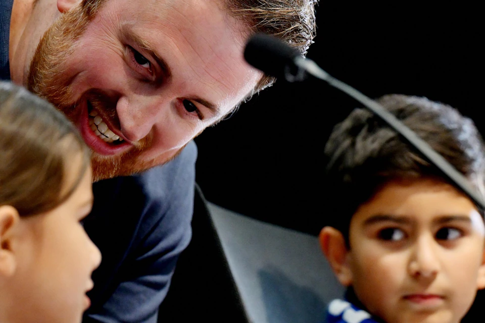 A man leans toward two children, engaging in conversation. The children are seated and one wears a blue and white scarf.