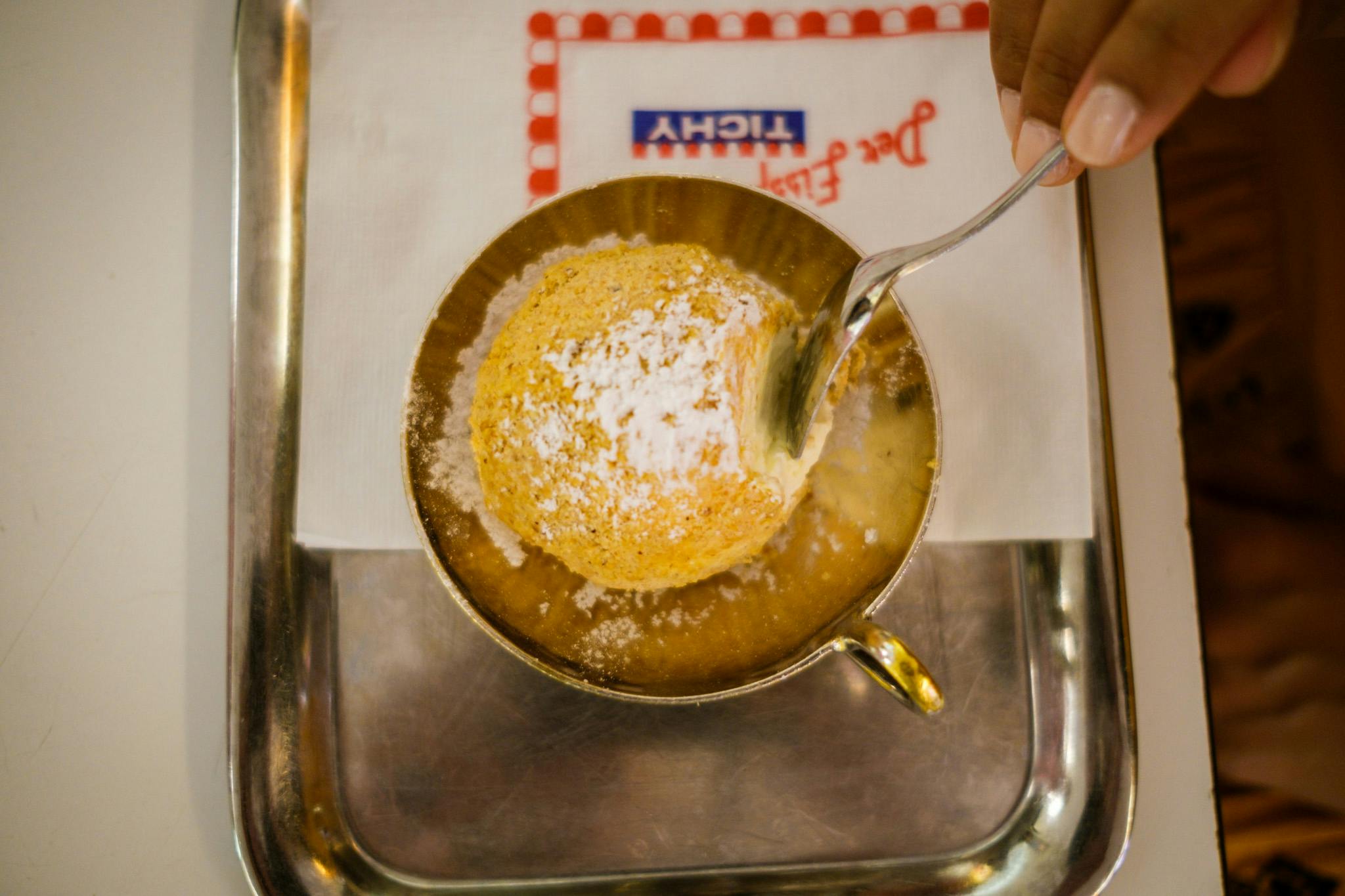 Osoba nabírající práškový dezert z šálku na stříbrném podnose s ubrouskem s modrým a červeným textem.