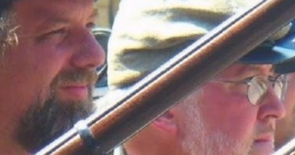 Two men in military uniforms and hats holding rifles, outdoors, facing the same direction.