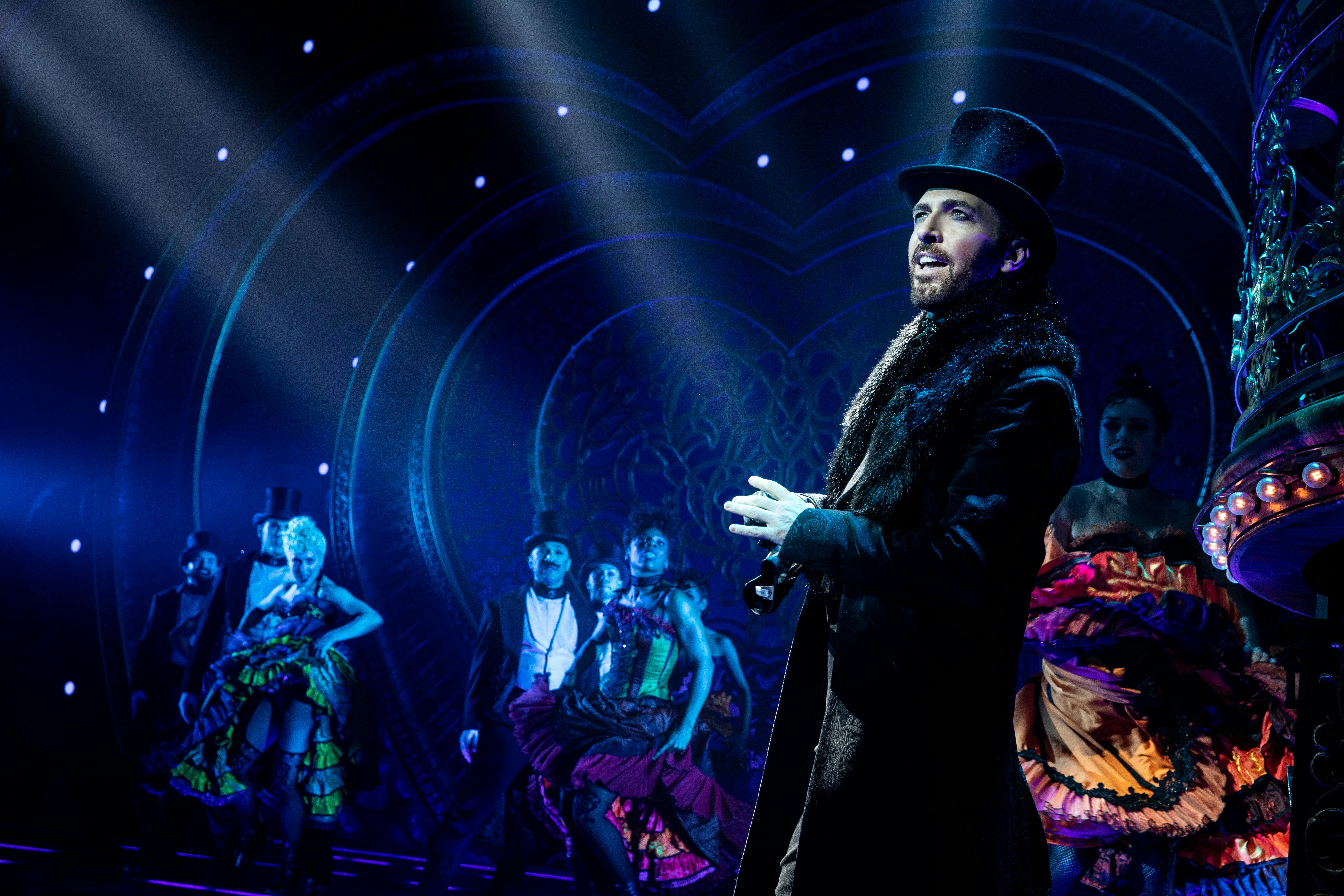A performer in a top hat and coat sings on stage, surrounded by dancers in colorful costumes under dramatic lighting.