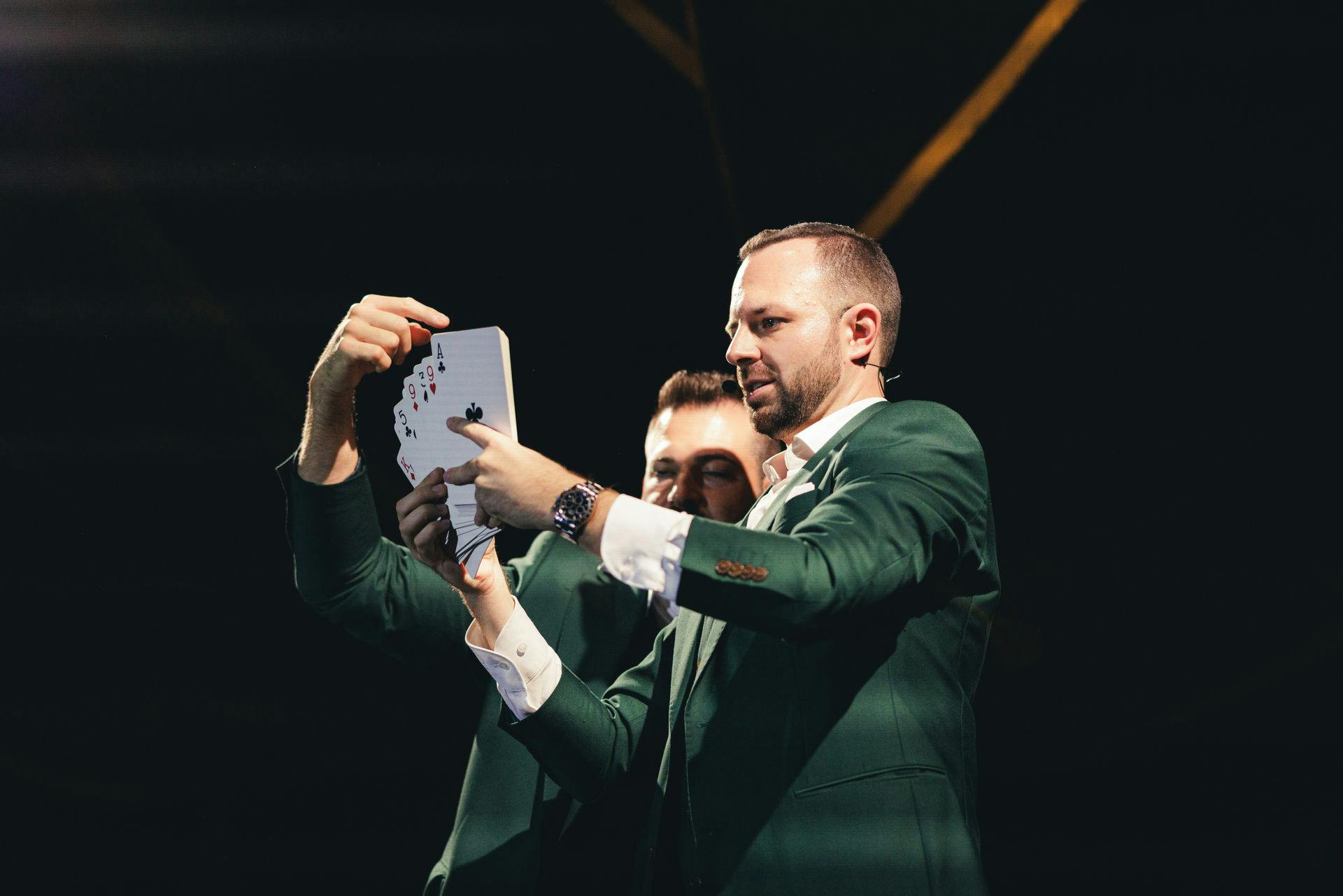 两名穿着绿色西装的男子在舞台灯光下表演卡牌戏法，重点是扑克牌迷。
