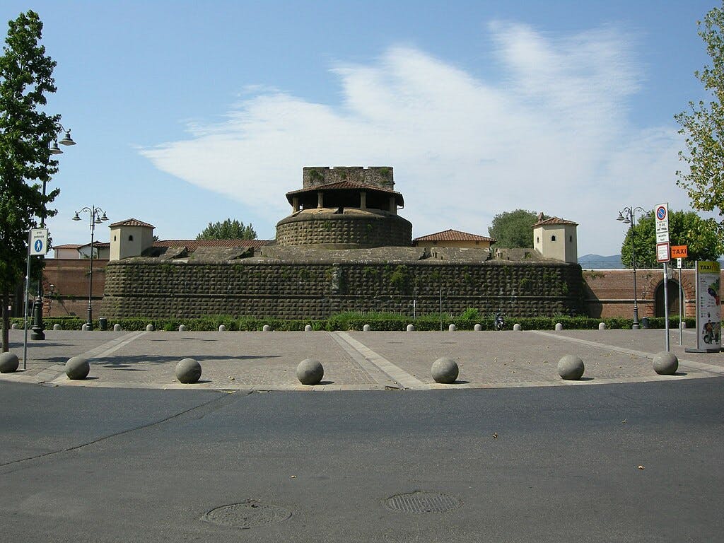 这座历史悠久的堡垒拥有厚厚的石墙，周围环绕着一片铺好的行人专用区、树木和路灯，周围多云。