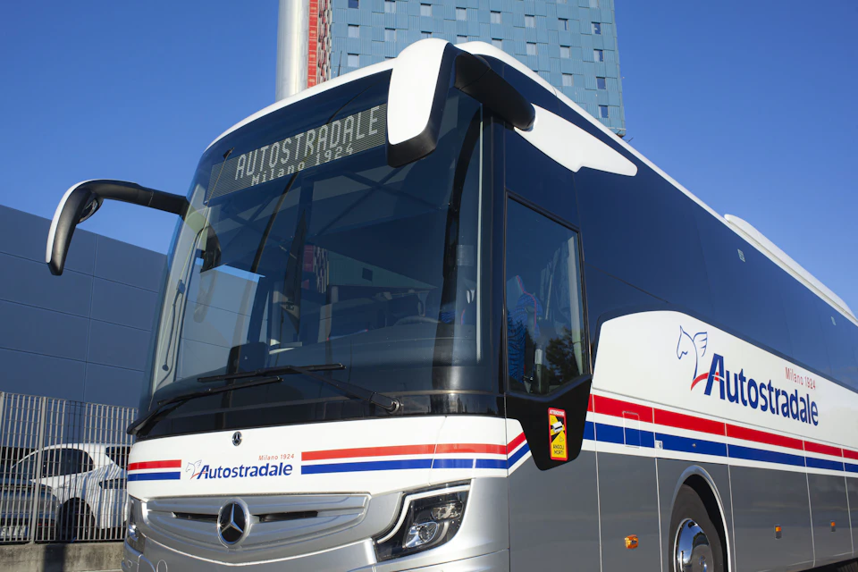 A modern Autostradale bus with "Milano 1924" displayed on its digital sign, parked in front of a tall, patterned building.