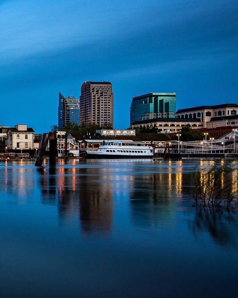 Panoráma města za soumraku s budovami, osvětlenou kavárnou a trajektem na klidných vodách, které odrážejí světla a modrou oblohu.
