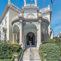 Nyklassisk byggnad med storslagen entré, flankerad av korintiska pelare, grekiska och europeiska flaggor och frodig grönska.