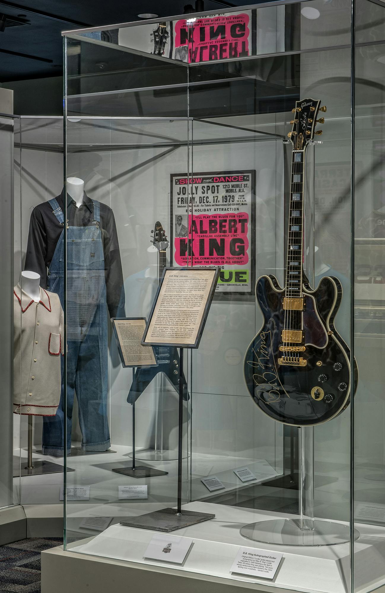 Display case with guitar, clothing, concert poster, and framed document. Items include black guitar, jumpsuit, shirt, and Albert King poster.