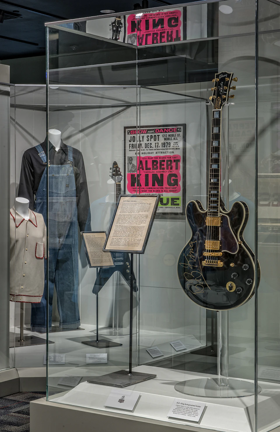 Display case with guitar, clothing, concert poster, and framed document. Items include black guitar, jumpsuit, shirt, and Albert King poster.