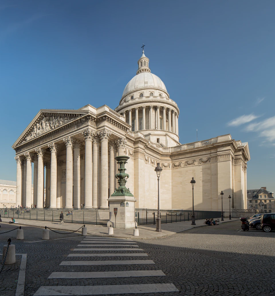 A neoclassical building with large columns and a dome under a clear blue sky, seen from a crosswalk on a cobblestone street.