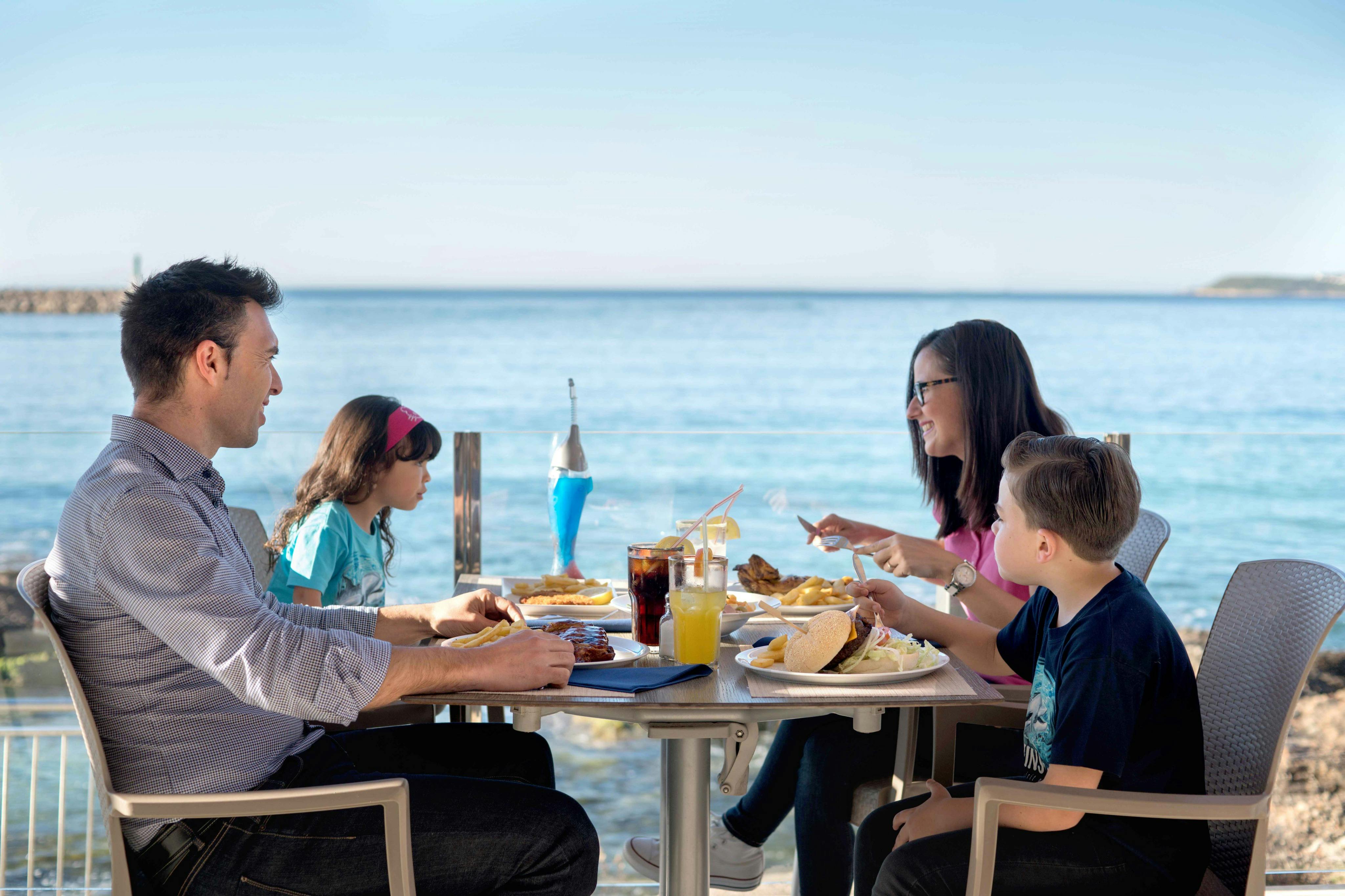 En familie, der spiser udendørs ved et bord tæt ved havet, med tallerkener med mad og drikkevarer på bordet under en klar blå himmel.