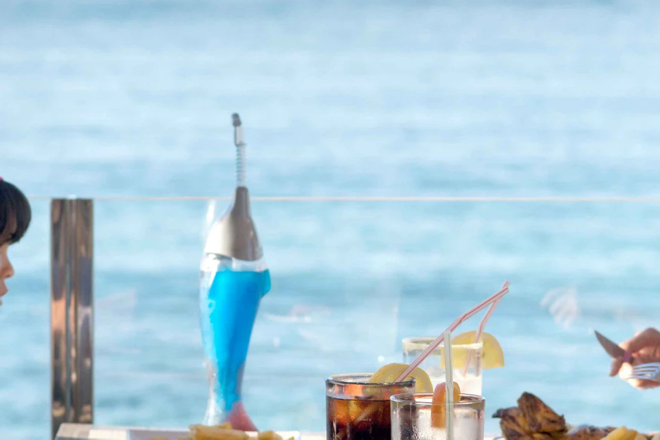 A family dining outdoors at a table near the sea, with plates of food and drinks on the table under a clear blue sky.