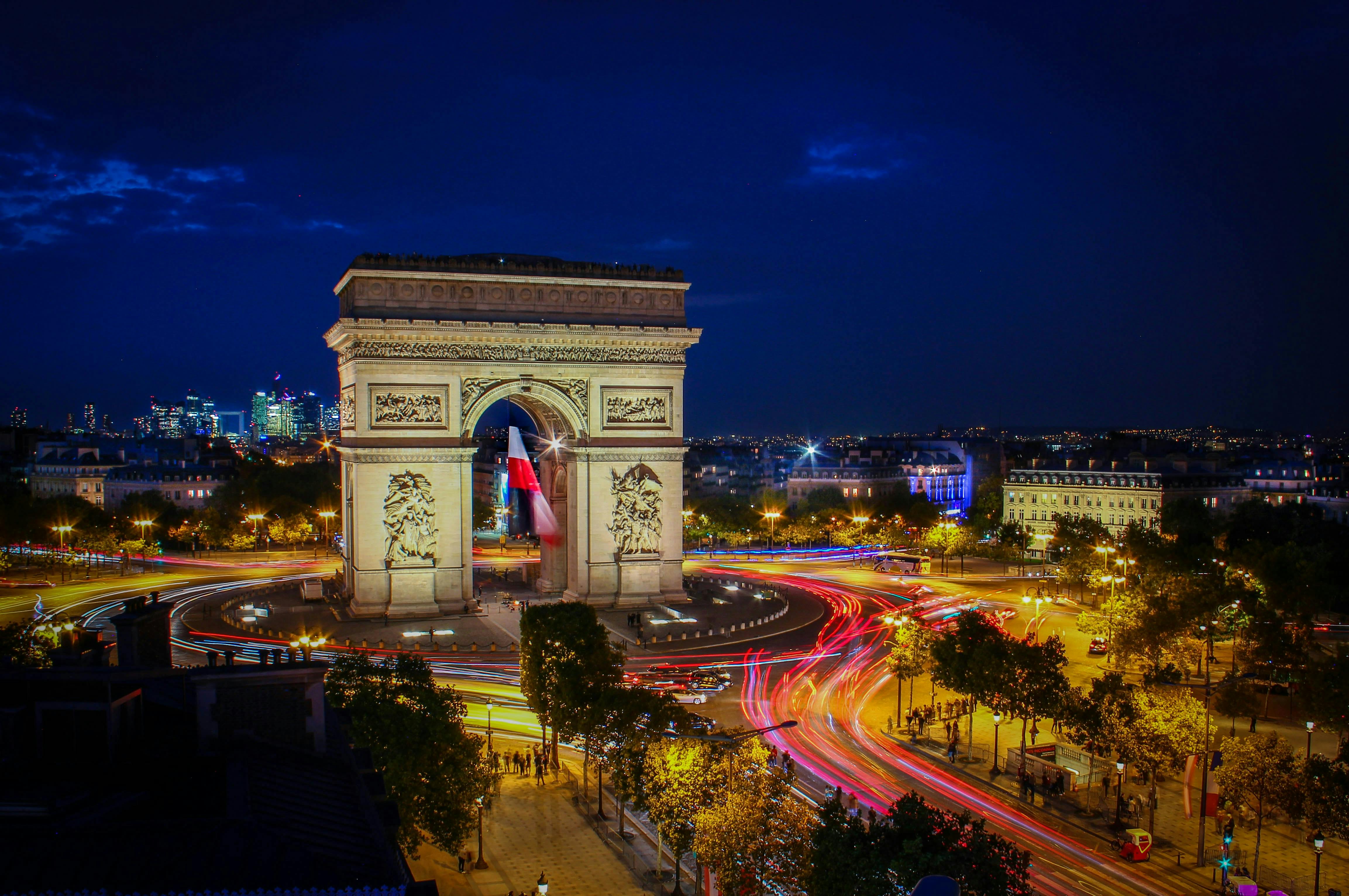 The Arc de Triomphe illuminated at night with light trails from traffic circling it and a cityscape in the background.