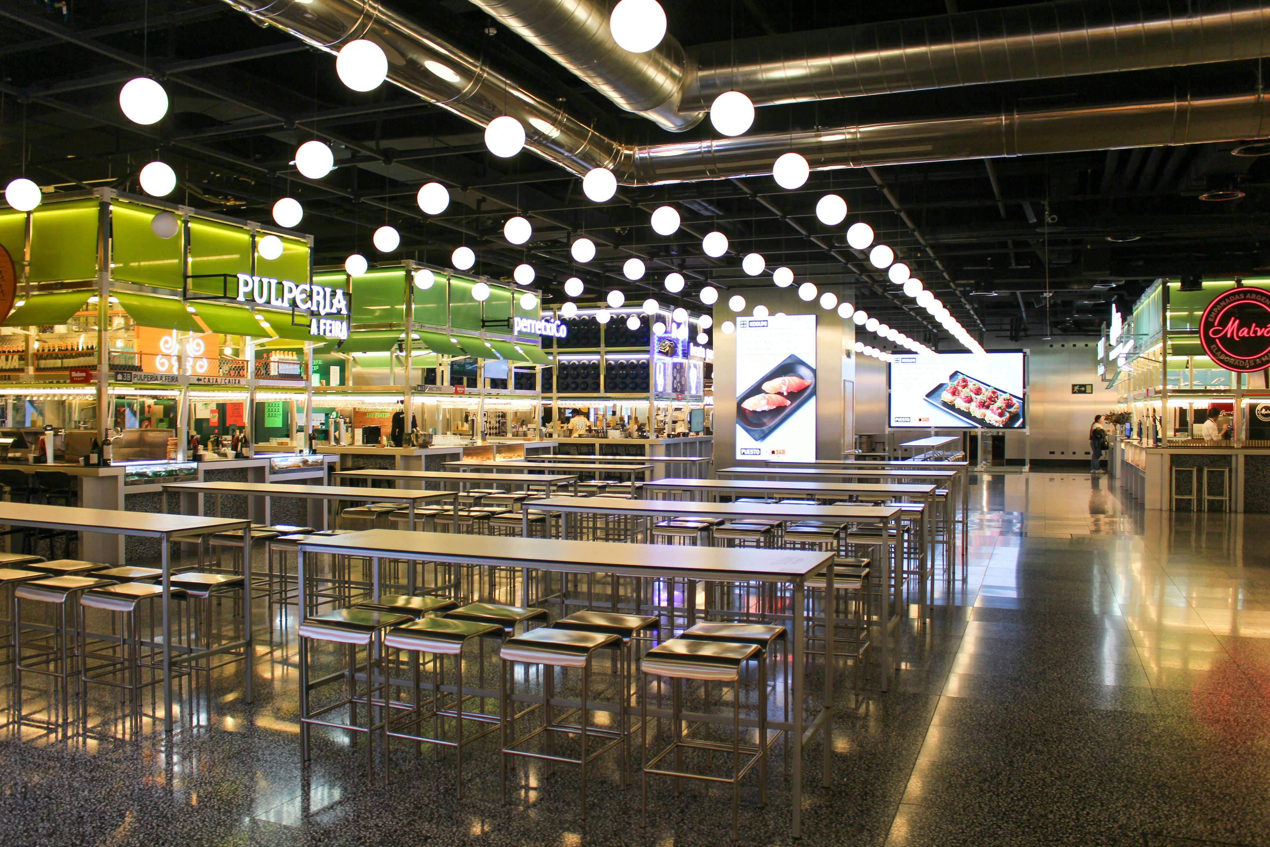 A modern food court with metal tables, stools, and bright lighting. Digital screens and various food stalls are visible.