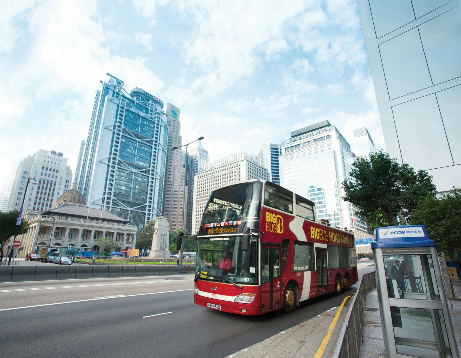 青空の下、背の高いモダンな建物に囲まれた街の通りを「Big Bus Hong Kong」の看板が付いた赤いダブルデッカーバスが走っています。