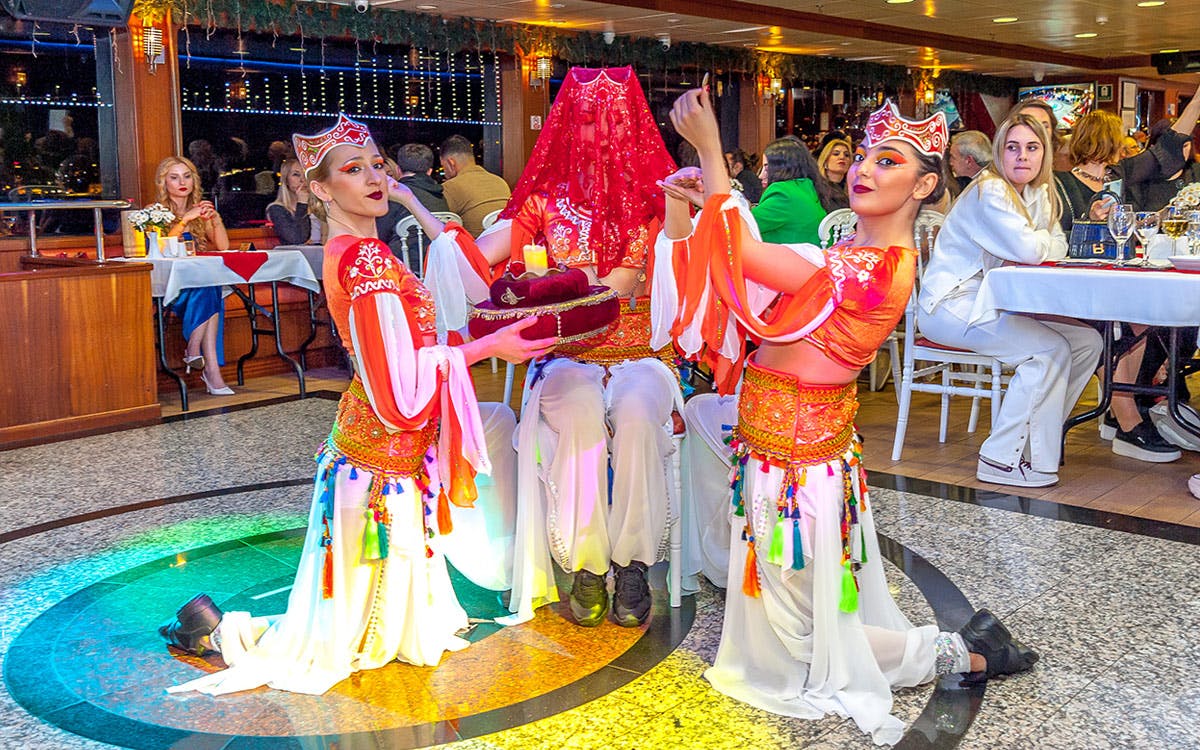 A dancer in traditional attire performs in a crowded restaurant, with another individual dressed similarly and a woman in white watching.