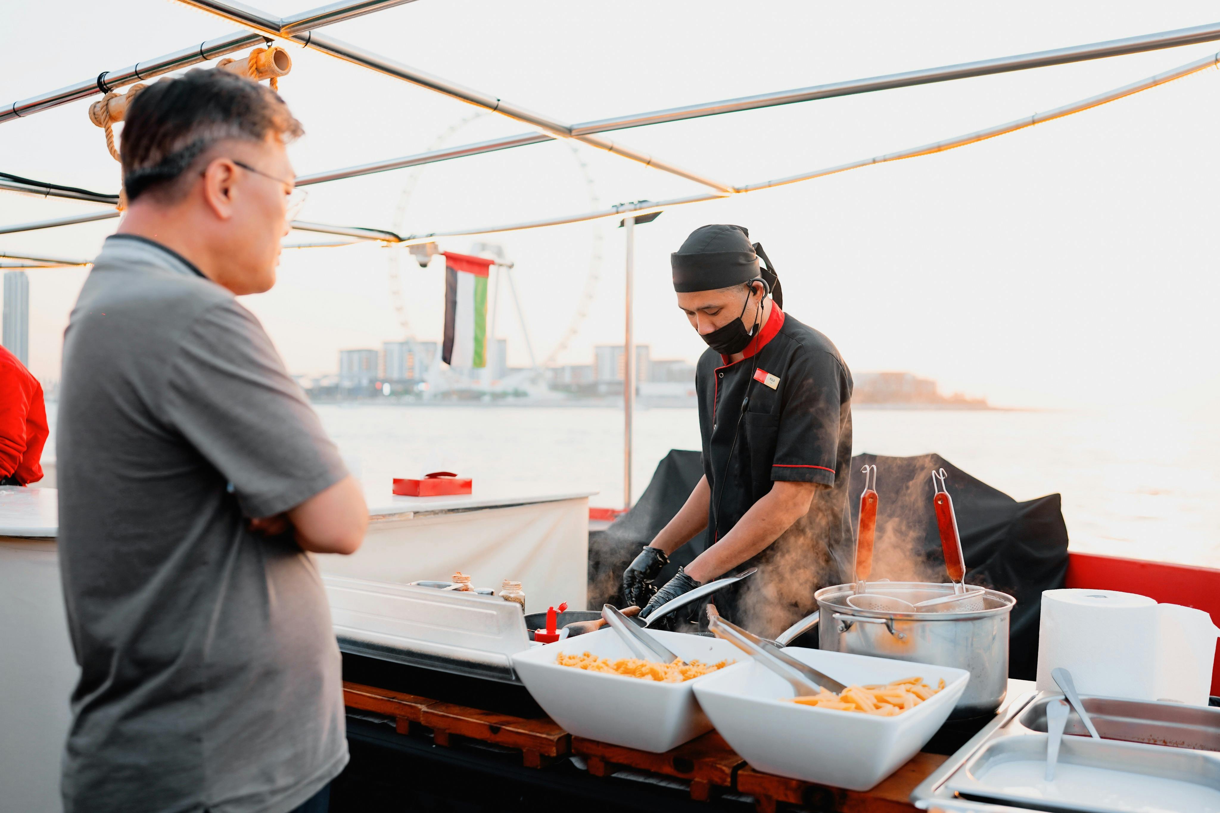 Šéfkuchař připravuje jídlo ve venkovním stánku, zatímco se zákazník dívá. Scéna obsahuje mísy s jídlem a v pozadí velké ruské kolo.