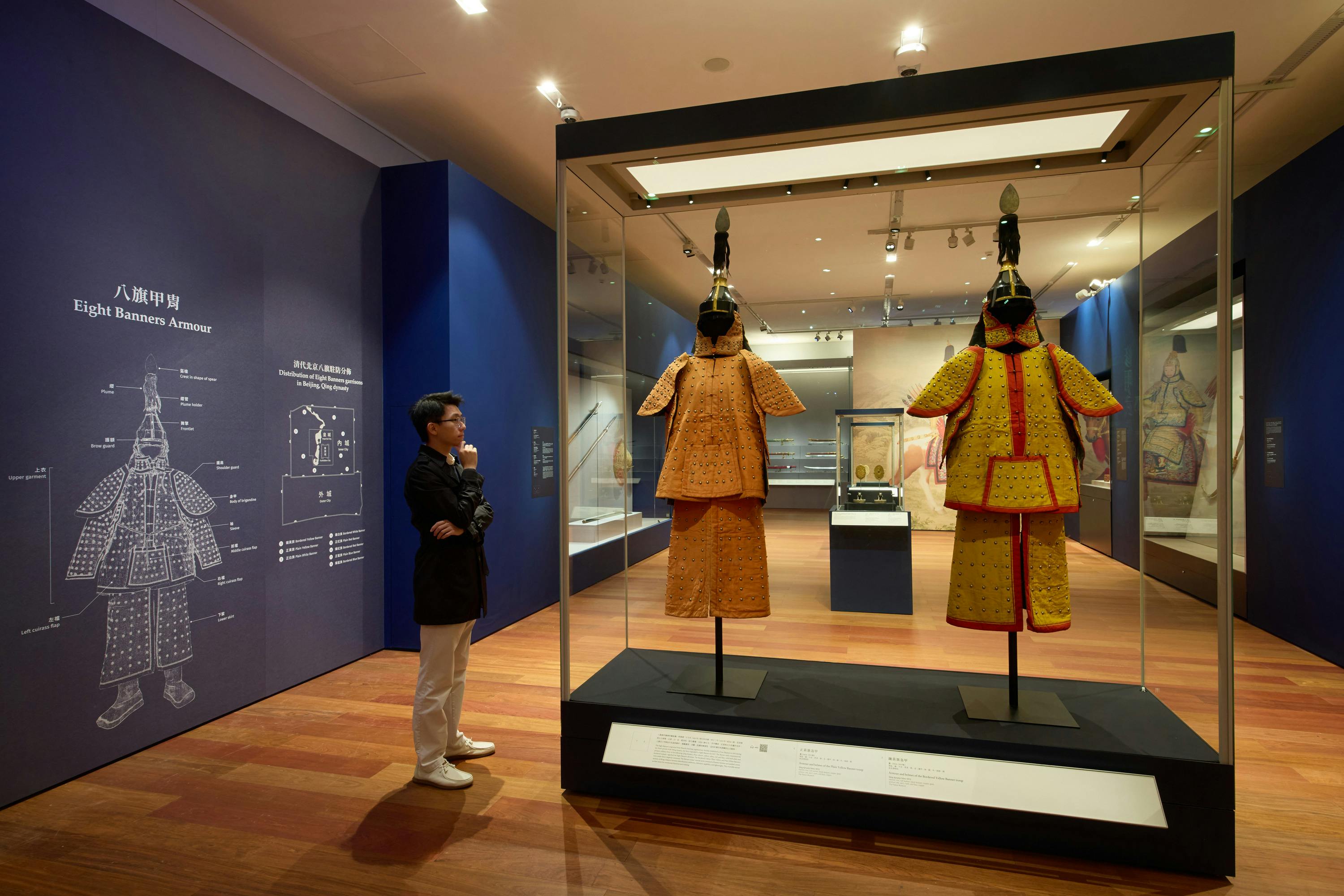 A person observes a traditional orange armor display in a museum, with educational diagrams on the wall nearby.