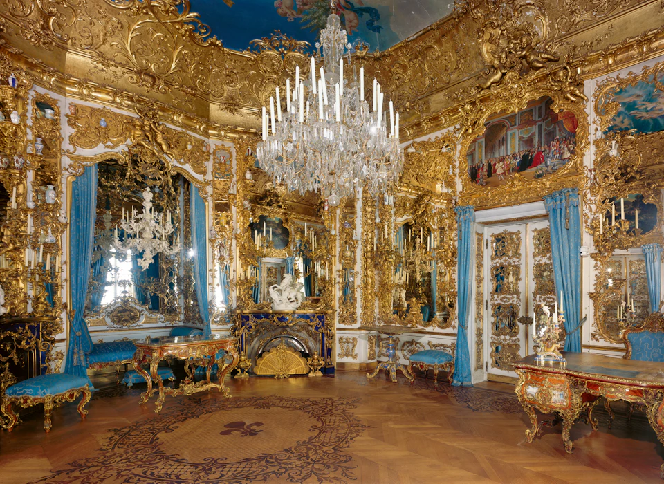 Ornate room with gold accents, grand chandelier, mirrors, intricate wood floor, blue drapes, decorative furniture, and wall art.