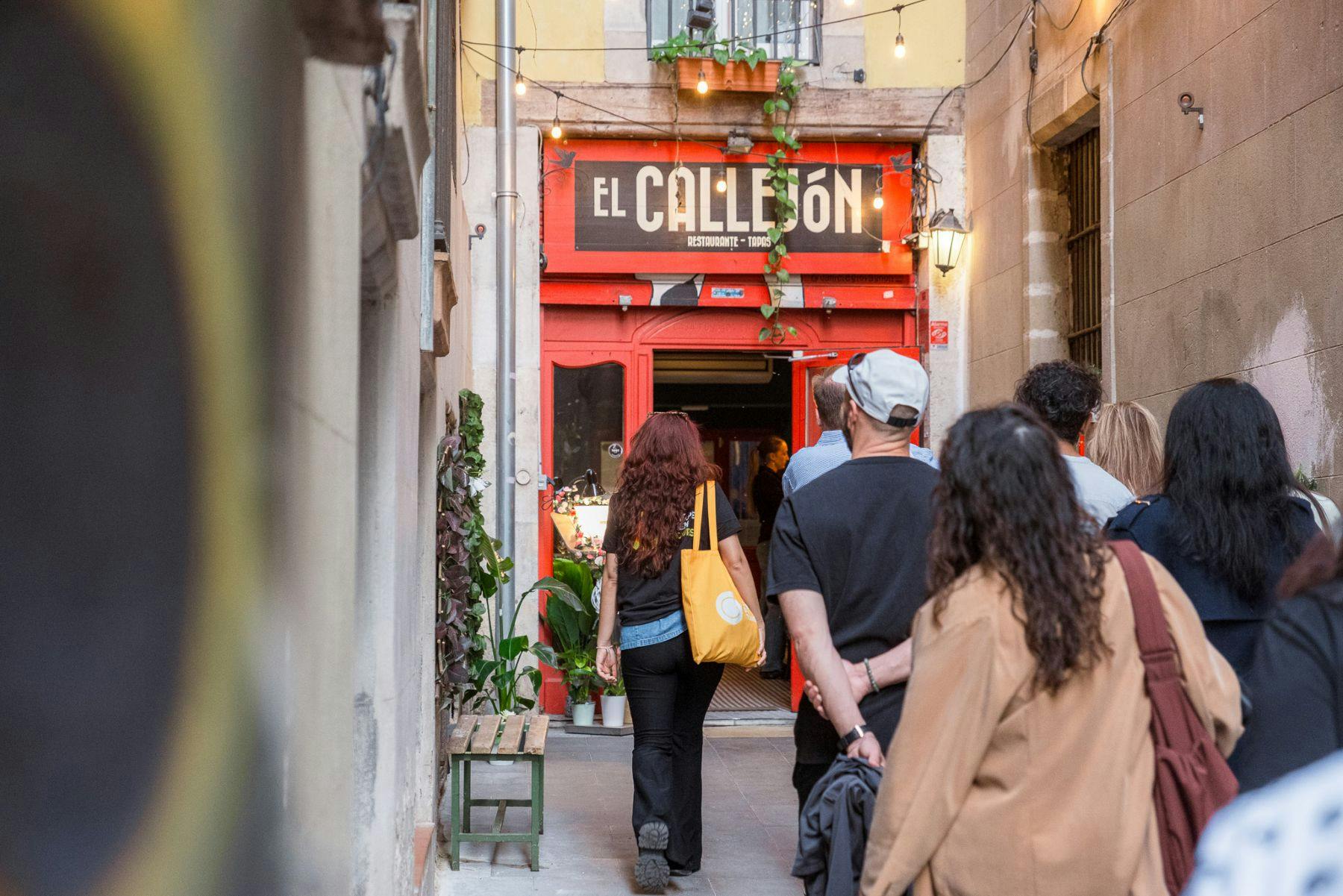 Pessoas a caminhar em direção à entrada de um restaurante com um letreiro que diz "El Callejón" numa ruela adornada com luzes e plantas.