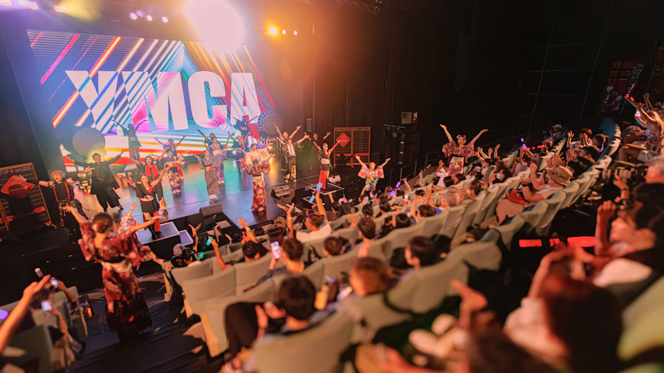 A vibrant stage performance with a band and dancers in traditional attire. Audience members enthusiastically participate.
