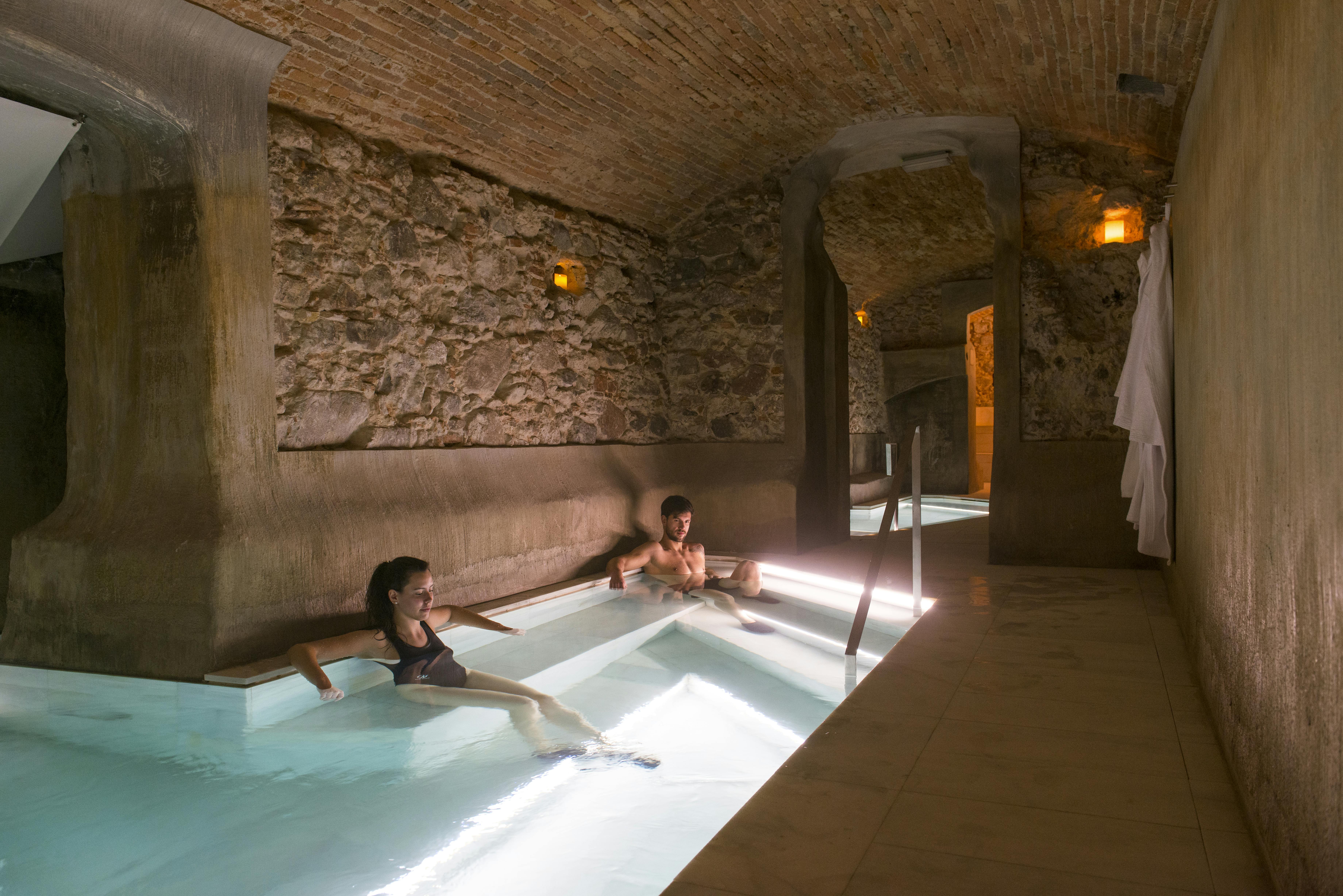 Two people relaxing in an indoor stone spa with warm lighting, reclined in shallow water under an arched brick ceiling.
