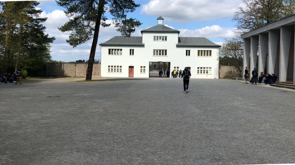 A person walking on a wide, empty cobblestone path toward a white building with a central clock tower and multiple windows.