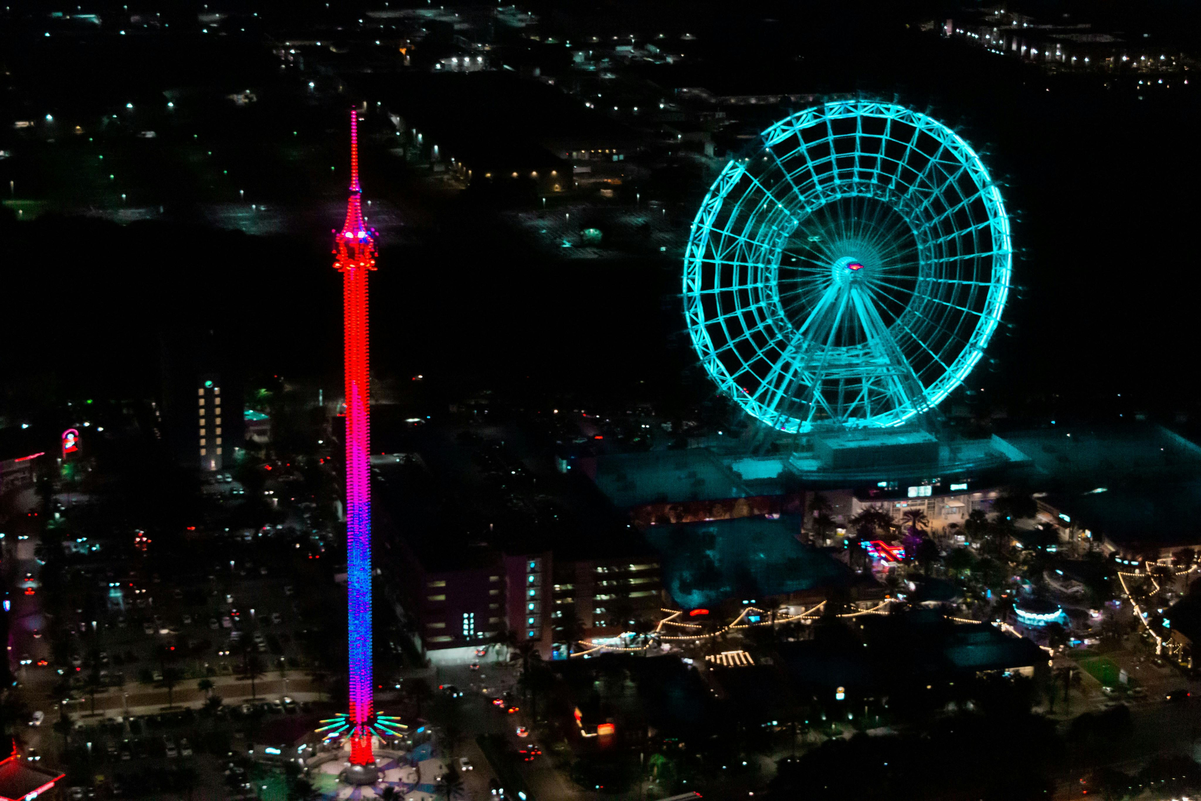 Nighttime view of an illuminated ferris wheel and drop tower ride in a busy amusement park.