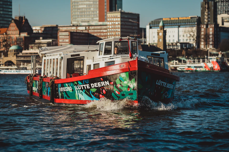 A colorful boat named "Lütte Deern" navigates through water with city buildings in the background under a clear sky.