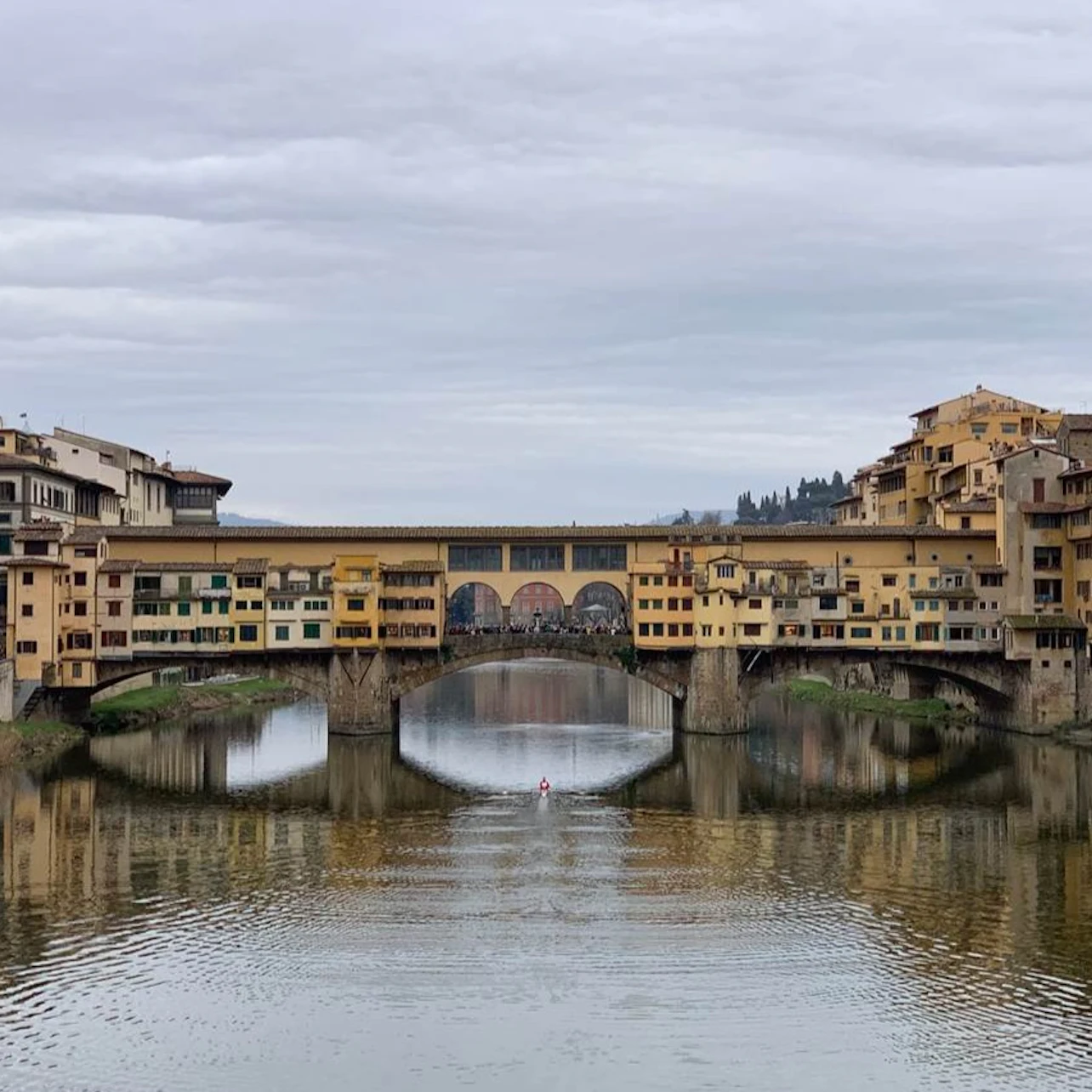 A historic bridge with shops spans a calm river, with reflections of the buildings and an overcast sky above.
